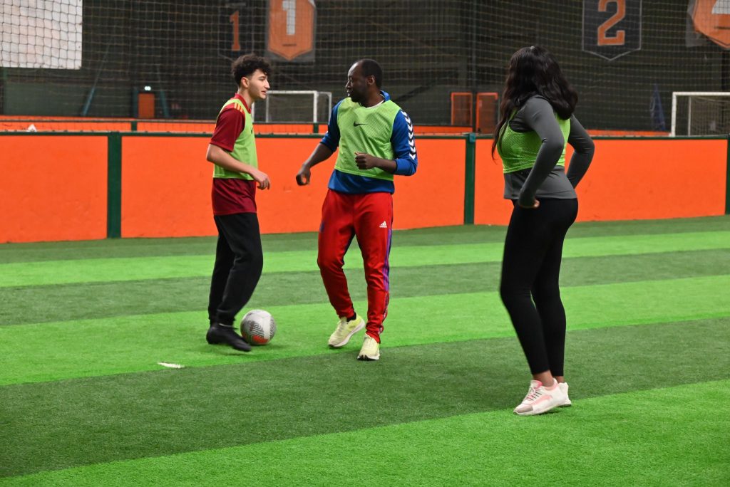 "Des collègues disputent un match de foot en entreprise sur un terrain en salle, portant des dossards aux couleurs vives. Un moment de convivialité et d’esprit d’équipe illustrant la cohésion entre collaborateurs.