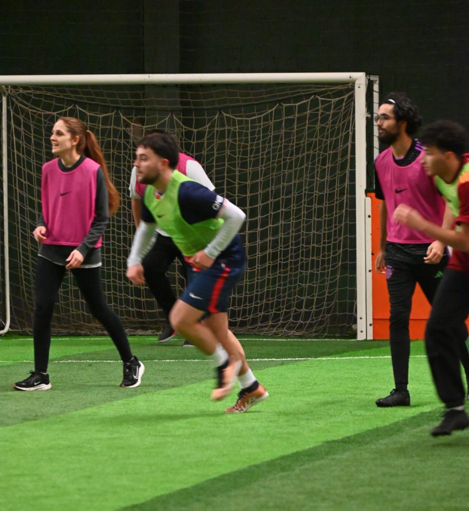 "Des collègues disputent un match de foot en entreprise sur un terrain en salle, portant des dossards aux couleurs vives. Un moment de convivialité et d’esprit d’équipe illustrant la cohésion entre collaborateurs.