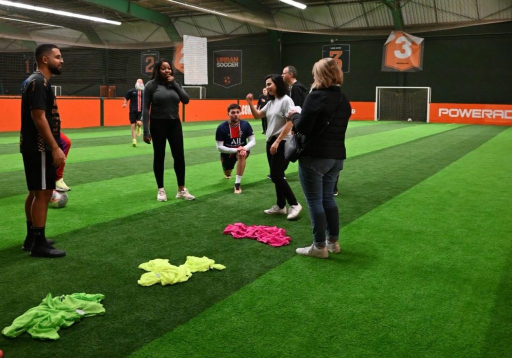 "Des collègues disputent un match de foot en entreprise sur un terrain en salle, portant des dossards aux couleurs vives. Un moment de convivialité et d’esprit d’équipe illustrant la cohésion entre collaborateurs.